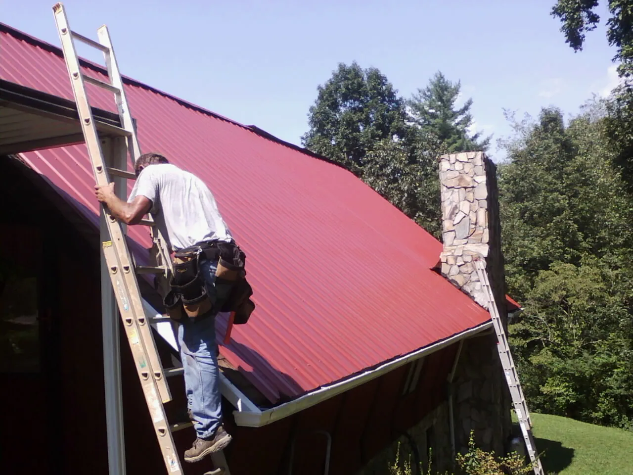 Roofer on a ladder beside a red metal roof and stone chimney — Soffit & Fascia Repair by Roofing Brothers in Kents Store